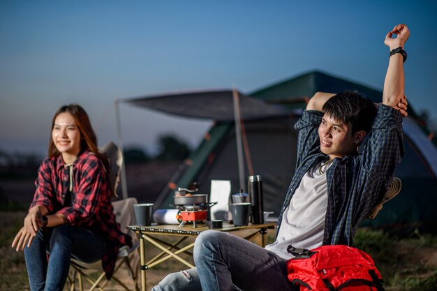 Selektiver Fokus Gut aussehender Mann, der auf einem Stuhl sitzt und die Arme in der Nähe seiner Freundin vor dem Campingzelt ausstreckt. Sie lächeln mit Glück und Frische, wenn sie sich in der Natur entspannen.