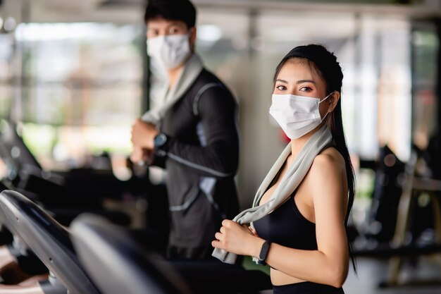 Selektiver Fokus, junge sexy Frau in Maske mit Sportbekleidung und Smartwatch und verschwommener junger Mann, Sie laufen auf dem Laufband, um im modernen Fitnessstudio zu trainieren