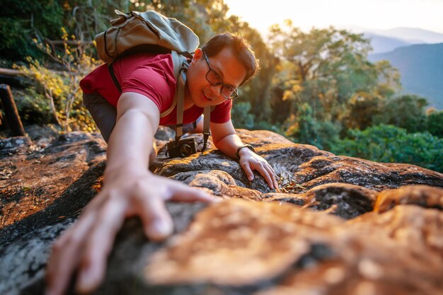 Selektiver Fokus Junger touristischer Rucksackmann mit Brille, der versucht, den Felsen hinaufzuklettern, um das Ziel zu erreichen Trekking-Reisekonzept Sonnenlicht hinter ihm Kopierraum