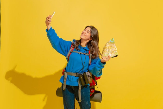 Selfie oder Vlog nehmen. Porträt eines fröhlichen jungen kaukasischen Touristenmädchens mit Tasche und Fernglas lokalisiert auf gelbem Studiohintergrund. Vorbereitung auf Reisen. Resort, menschliche Gefühle, Urlaub.