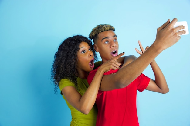 Selfie oder Vlog zusammen nehmen. Junger emotionaler afroamerikanischer Mann und Frau in den bunten Kleidern auf blauem Hintergrund. Schönes Paar. Konzept der menschlichen Emotionen, Gesichtsausdruck, Beziehungen, Anzeige.