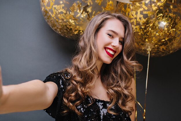 Selfie-Porträt der jungen charmanten Frau mit den roten Lippen, dem langen brünetten Haar, das mit großen Luftballons voll mit goldenen Lametta lächelt. Positivität ausdrücken, Party feiern.