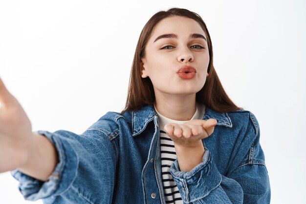 Senden von Küssen Nette junge Frau, die selfie nimmt, sendet während der Videoanrufkonferenz, die über weißem Hintergrund steht, einen Luftkuss in die Kamera