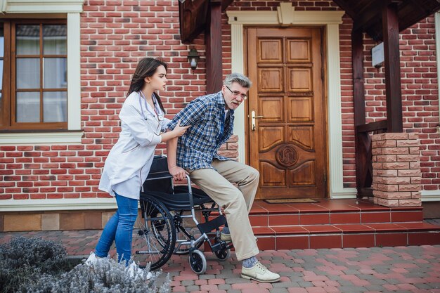 Senior Mann sitzt im Rollstuhl mit lächelnder Krankenschwester, kümmert sich um und diskutiert und jubelt im Garten im Pflegeheim