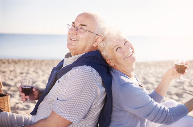 Senior sitzt Rücken an Rücken und trinkt Wein. Älteres Paar im Strand-, Ruhestands- und Sommerferienkonzept