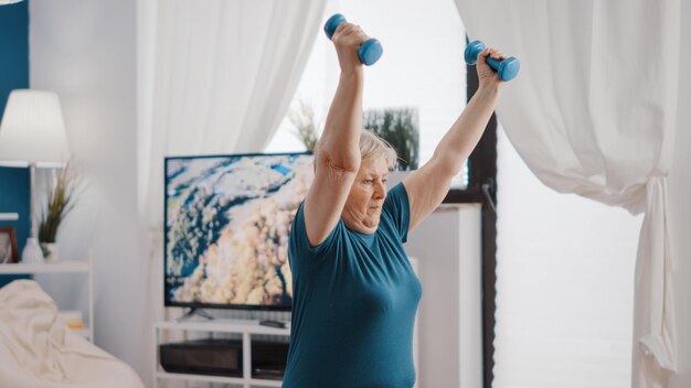 Seniorentraining mit Hanteln und Sitzen auf dem Toningball, um auf der Yogamatte zu trainieren und zu trainieren. Ältere Frau, die Gewichte hebt und die Armmuskulatur streckt, um für Wellness zu trainieren.