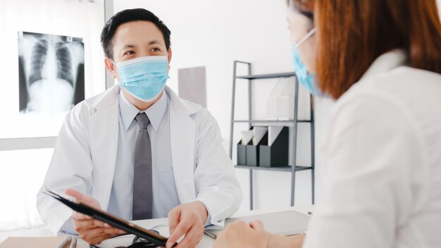 Seriöser asiatischer männlicher Arzt, der eine Schutzmaske mit Tablet trägt, liefert großartige Nachrichten und diskutiert die Ergebnisse