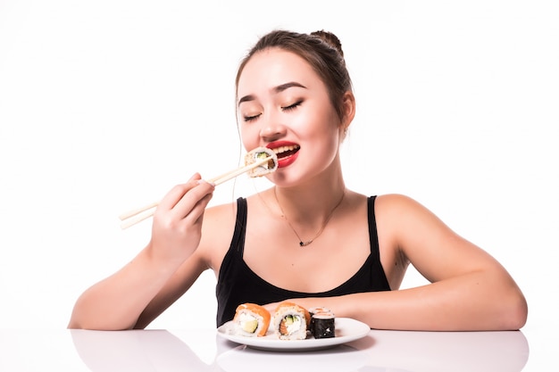Sexy asiatischen Blick mit bescheidenen Frisur sitzen auf dem Tisch essen Sushi-Rollen lächelnd isoliert auf weiß