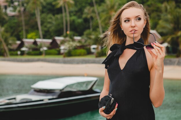 Sexy attraktive Luxusfrau gekleidet im schwarzen Kleid, das auf Pier im Luxusresorthotel aufwirft, Sonnenbrille tragend, Sommerferien, tropischer Strand