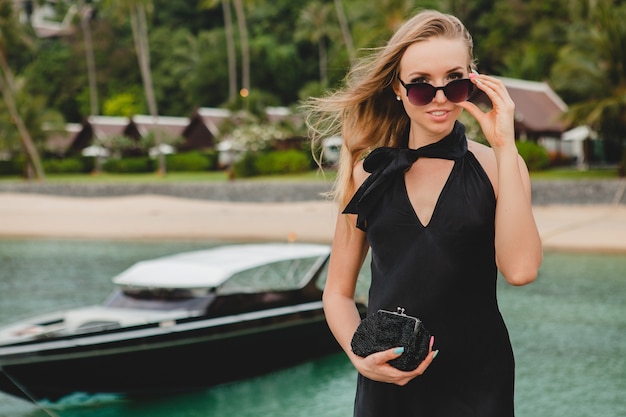 Sexy attraktive Luxusfrau gekleidet im schwarzen Kleid, das auf Pier im Luxusresorthotel aufwirft, Sonnenbrille tragend, Sommerferien, tropischer Strand