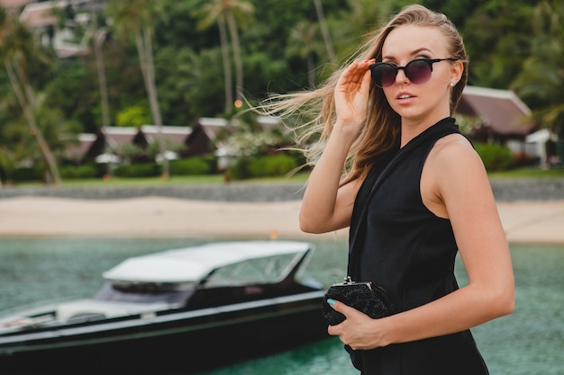 Sexy attraktive Luxusfrau gekleidet im schwarzen Kleid, das auf Pier im Luxusresorthotel aufwirft, Sonnenbrille tragend, Sommerferien, tropischer Strand