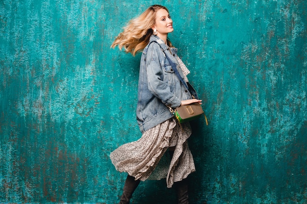 Kostenloses Foto sexy attraktive stilvolle blonde frau in jeans und übergroße jacke, die gegen vintage grüne wand in der straße geht