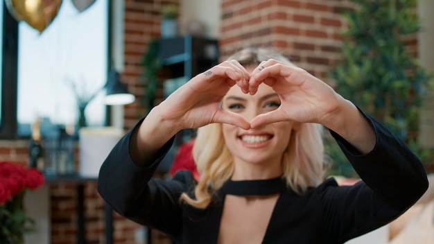 Sexy blonde Frau zeigt herzförmiges Zeichen vor der Kamera, um den Valentinstag mit Blumen und Geschenken zu feiern. Attraktive schöne Person, die Romantik und Liebessymbol bewirbt.