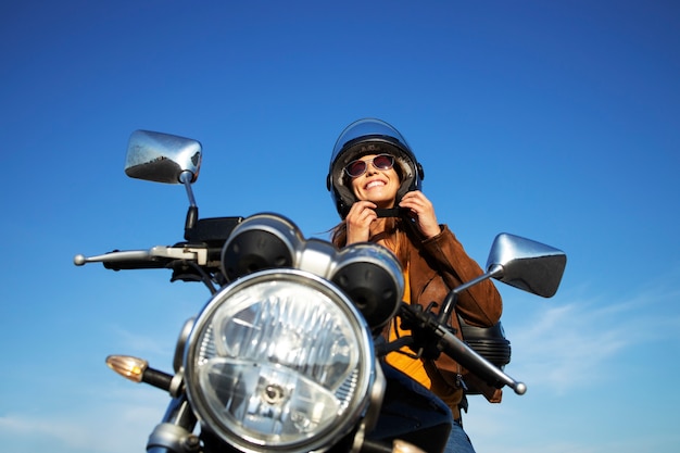 Sexy brünette Frau in der Lederjacke, die Helm aufsetzt und auf Retro-Artmotorrad an schönem sonnigem Tag sitzt