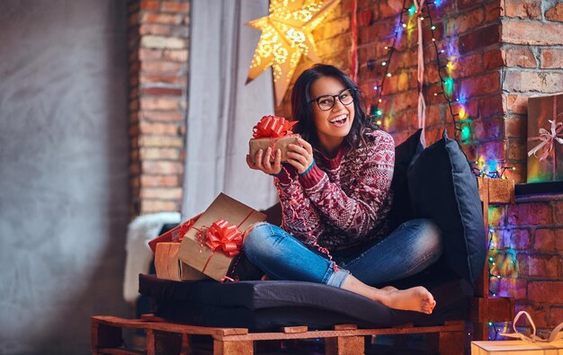 Sexy brünette Frau mit nacktem Fuß in Jeans und rotem Pullover posiert auf einem Sofa in einem Raum mit Weihnachtsdekoration.