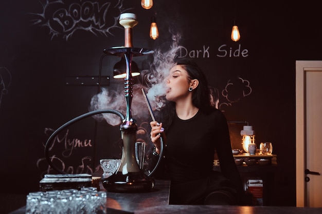 Sexy brünettes Mädchen in verführerischer schwarzer Kleidung raucht eine Wasserpfeife, während sie in einem Nachtclub auf der Theke sitzt.
