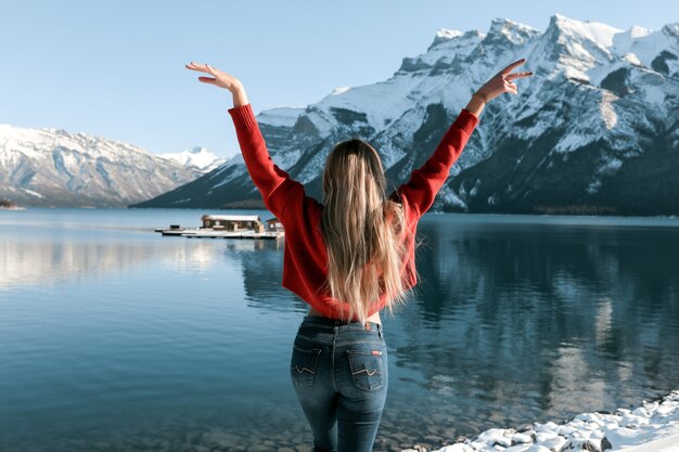 Sexy Dame mit schlankem perfektem Körper, der am Strand nahe dem Wintersee steht. Weißer Schnee liegt auf dem Boden und auf den Gipfeln der Berge. Langes blondes Haar, das auf der Rückseite des roten Pullovers liegt.