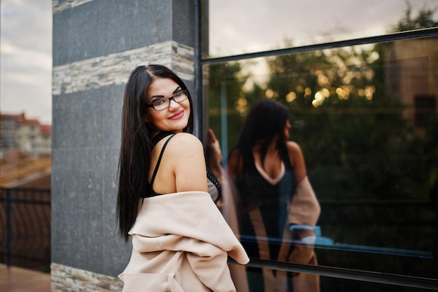 Sexy Frau mit schwarzen Haaren in Brille und Mantel posierte vor einem Gebäude mit modernen Fenstern