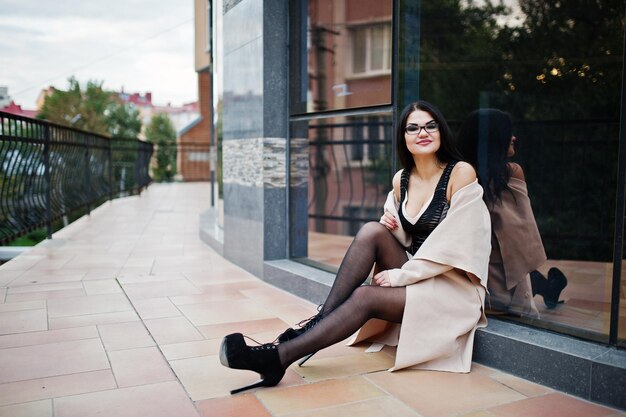 Sexy Frau mit schwarzen Haaren in Brille und Mantel posierte vor einem Gebäude mit modernen Fenstern