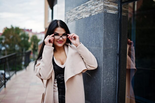 Sexy Frau mit schwarzen Haaren in Brille und Mantel posierte vor einem Gebäude mit modernen Fenstern