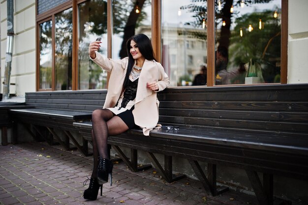 Sexy Frau mit schwarzen Haaren in Brille und Mantel sitzt auf einer Bank mit Handy zur Hand und macht Selfie