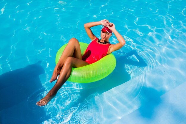 Sexy gebräunte Frau im roten Badeanzug, die Spaß hat und den Sommer in einem erstaunlich großen Swimmingpool genießt Junges hübsches Mädchen, das auf einem aufblasbaren Ring schwimmt Stilvolle transparente Kappe Strandparty