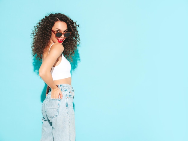 Sexy junge schöne lächelnde Frau in trendiger Sommer-Hipster-Kleidung sorglose Frau, die in der Nähe der blauen Wand im Studio posiertPositives brünettes Modell mit Afro-Locken-FrisurFröhlich und glücklich In Sonnenbrille