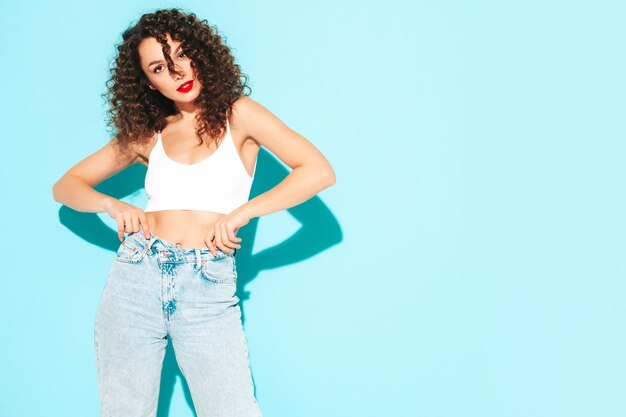 Sexy junge schöne lächelnde Frau in trendiger Sommer-Hipster-Kleidung sorglose Frau posiert in der Nähe der blauen Wand im Studio Positives brünettes Modell mit Afro-Locken-Frisur Fröhlich und glücklich