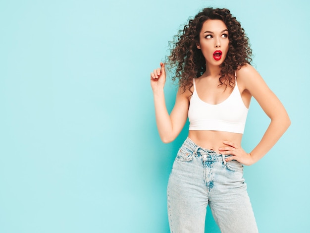 Sexy junge schöne lächelnde Frau in trendiger Sommer-Hipster-Kleidung sorglose Frau posiert in der Nähe der blauen Wand im Studio Positives brünettes Modell mit Afro-Locken-Frisur Fröhlich und glücklich