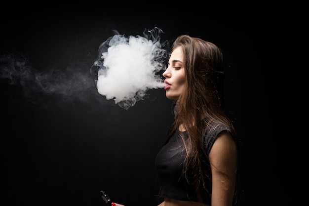 Sexy Mädchen in einem schwarzen Kleid, das elektronische Zigarette auf dunkler Wand raucht