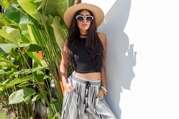 Sexy Model in Sonnenbrillen, die über grünen tropischen Pflanzen und weißer Wand aufwerfen. Trägt ein stilvolles Sommeroutfit, einen Strohhut und Accessoires.