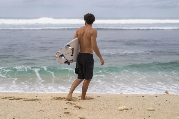 Sexy Surfer, der Mann mit Surfbrett surft. Hübscher junger männlicher Athlet, der Surfbrett mit nassen Haaren im Sommerstrandsporturlaub hält. Reiseziel für Sportreisen. Surfer-Lebensstil.
