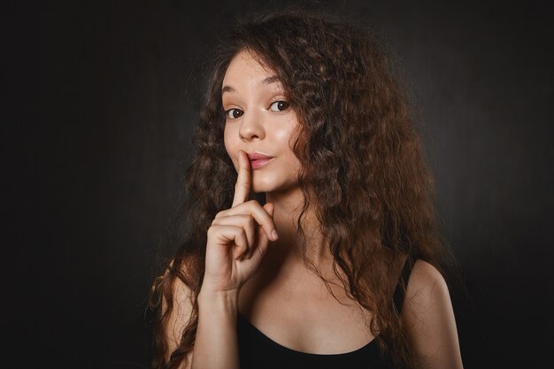 Shh. Erzähl es niemandem. Bild des emotionalen lustigen brünetten Mädchens im Trägershirt, das Vorder Ingwer an ihren Lippen hält, Augenbrauen hochzieht, Shh sagt und bittet, ihr Geheimnis nicht preiszugeben. Vertrauliche Informationen