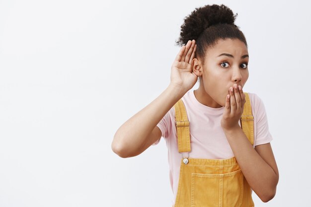 Shh, ich kann kein Flüstern hören. Faszinierte und verblüffte stilvolle Afroamerikanerin in gelben Overalls, mit Handfläche in der Nähe des Ohrs, Abhören oder belauschender schockierender Unterhaltung