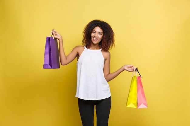 Shopping-Konzept - Close up Portrait junge schöne attraktive afrikanische Frau lächelnd und freudig mit bunten Einkaufstasche. Gelbe Pastellwand Hintergrund. Text kopieren
