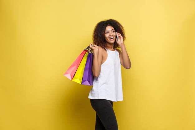 Shopping-Konzept - Close up Portrait junge schöne attraktive afrikanische Frau lächelnd und freudig mit bunten Einkaufstasche. Gelbe Pastellwand Hintergrund. Text kopieren