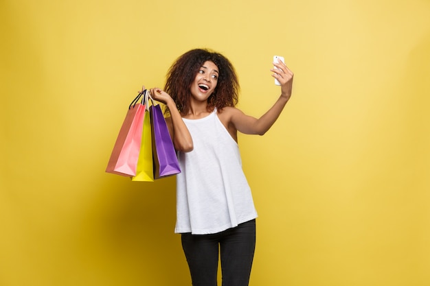 Shopping-Konzept - Close up Portrait junge schöne attraktive afrikanische Frau lächelnd und freudig mit bunten Einkaufstasche. Gelbe Pastellwand Hintergrund. Text kopieren