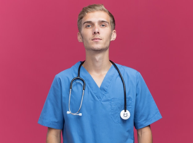Sicheres Betrachten der Kamera junger männlicher Arzt, der Arztuniform mit Stethoskop lokalisiert auf rosa Wand mit Kopienraum trägt