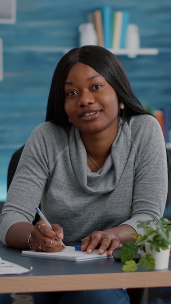 Sicht des Studenten mit dunkler Haut, der während des Kommunikations-Videoanruf-Telearbeits-Konferenztreffens in die Kamera lächelt. Afroamerikanische Frau, die am Schreibtisch im Wohnzimmer sitzt und von zu Hause aus arbeitet