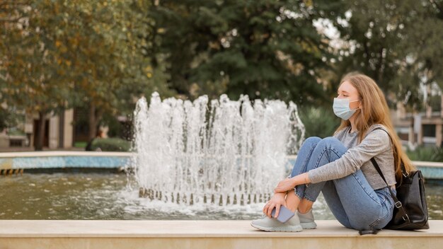 Sied Ansicht Frau, die eine medizinische Maske trägt, während sie neben einem Brunnen sitzt
