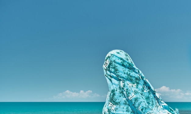 Silhouette einer Frau, die in einen Schal gehüllt ist, gegen einen Windstoß vor dem Hintergrund des Sommermeeres und des blauen, klaren Himmels, Wochenende auf See, kühle Brise