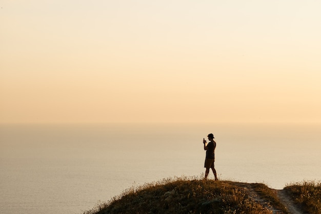 Silhouette eines jungen Mannes, der bei Sonnenuntergang auf einem Smartphone Fotos vom Meer macht. Abend, Sommerreise in den Urlaub