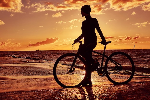 Silhouette eines Mädchens mit einem Fahrrad an der Küste bei Sonnenuntergang.