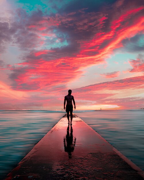 Silhouette eines Mannes, der auf einem Steinpier mit seinem Spiegelbild und den schönen atemberaubenden Wolken geht