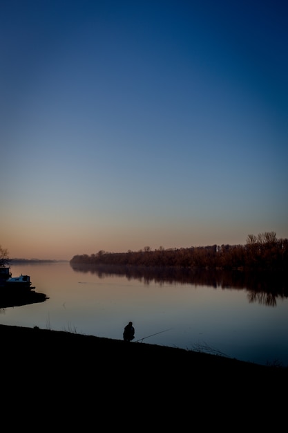 Silhouette eines Mannes, der nahe dem Wasser unter einem klaren blauen Himmel in einem vertikalen Schuss sitzt