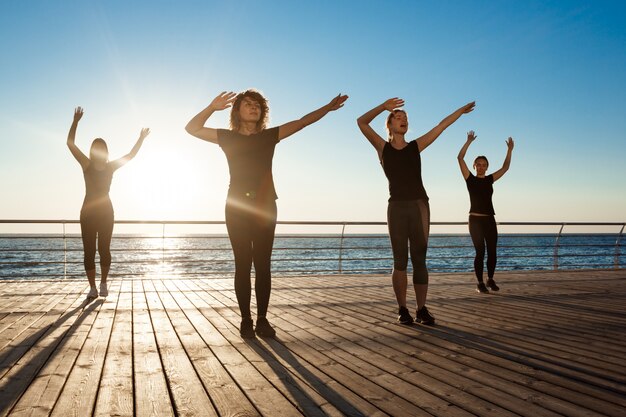 Silhouetten von sportlichen Frauen, die zumba nahe Meer bei Sonnenaufgang tanzen