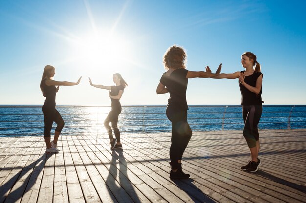 Silhouetten von sportlichen Frauen, die zumba nahe Meer bei Sonnenaufgang tanzen