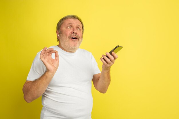 Singen mit Kopfhörer und Smartphone. Kaukasisches Mannporträt auf gelbem Studiohintergrund. Schönes männliches Modell im weißen Hemd.