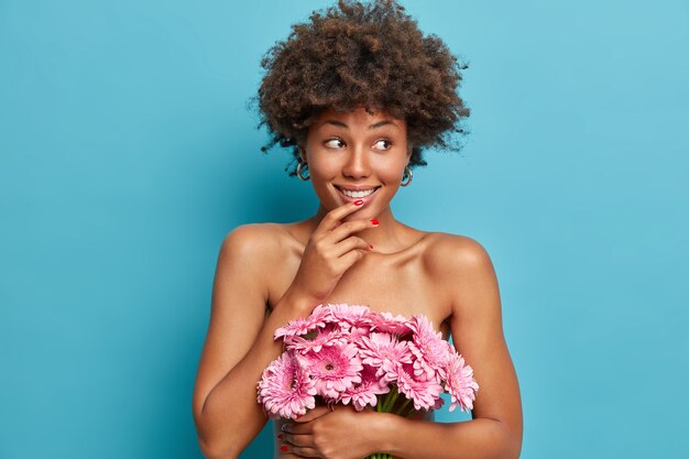 Sinnlich glückliches weibliches Modell mit gesundem nacktem Körper, hält Strauß von rosa Gerberablumen, schaut mit verträumtem fröhlichem Ausdruck beiseite, steht
