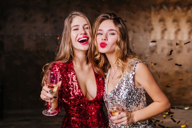 Sinnliche Frau im roten trendigen Kleid glücklich lachen, während ihre Freundin mit küssendem Gesichtsausdruck posiert